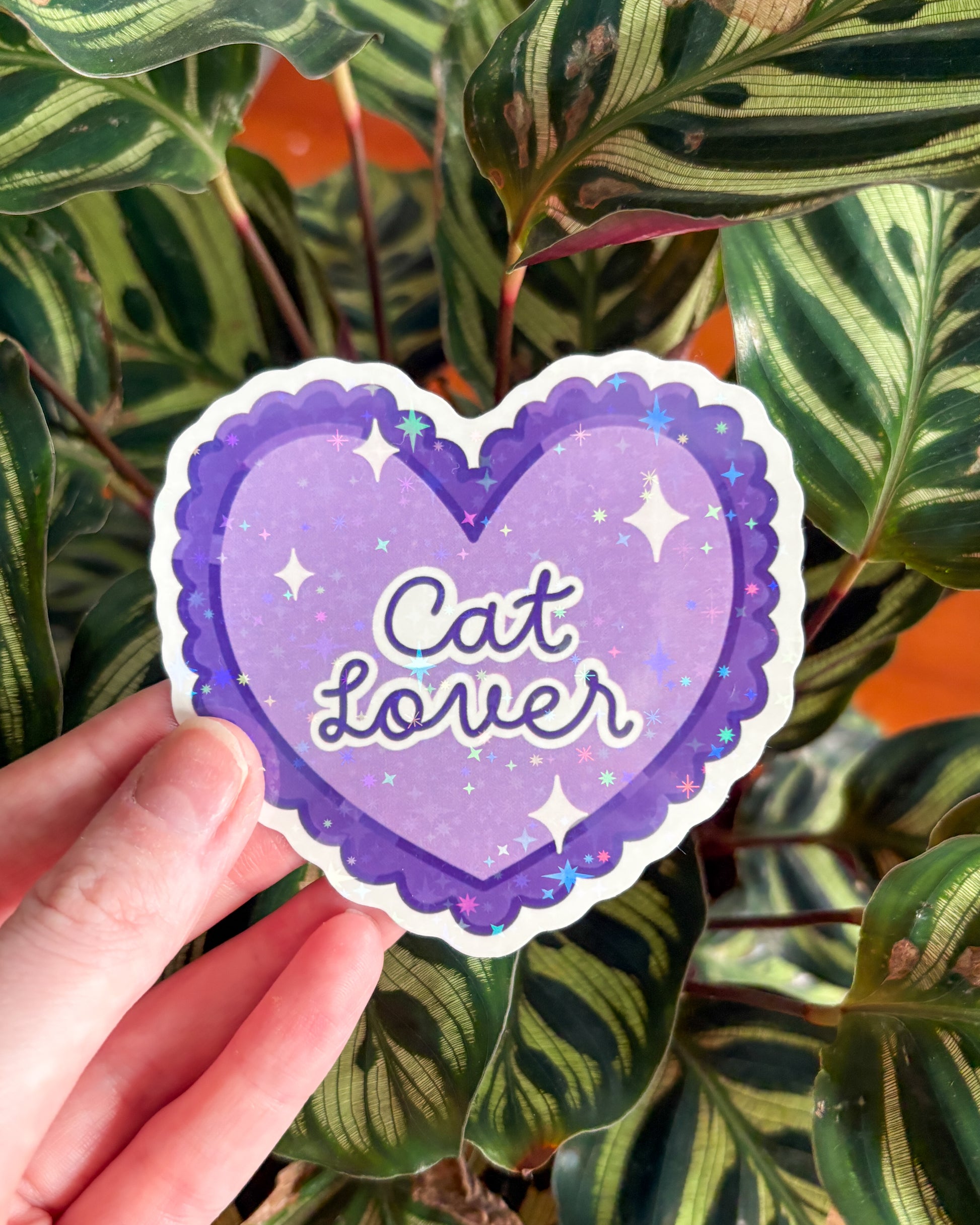 Heart-shaped sticker with 'Cat Lover' text held in front of green leaves
