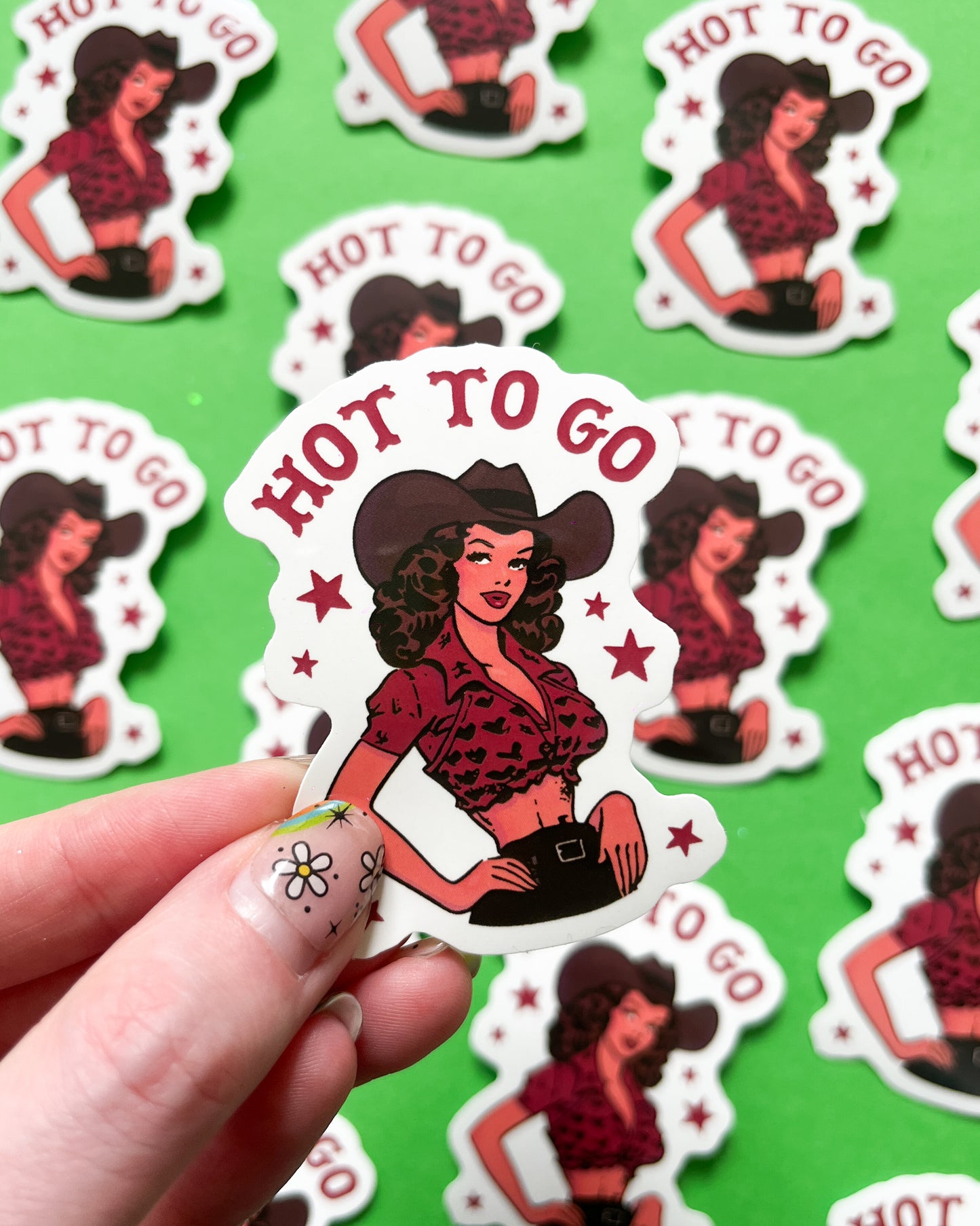 A person holding a vinyl sticker with an illustration of a woman in a cowgirl outfit, with the text 'HOT TO GO'.