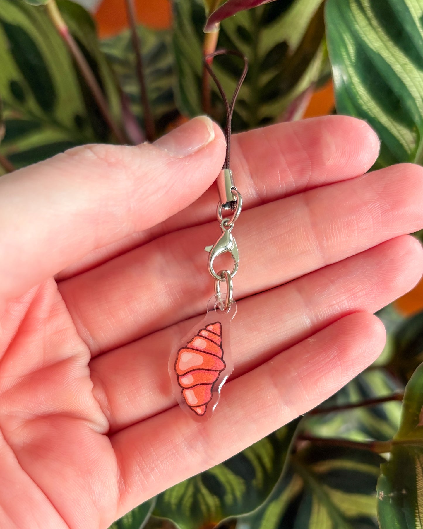 Hand holding a small orange and clear croissant charm with a clasp against a blurred plant background.