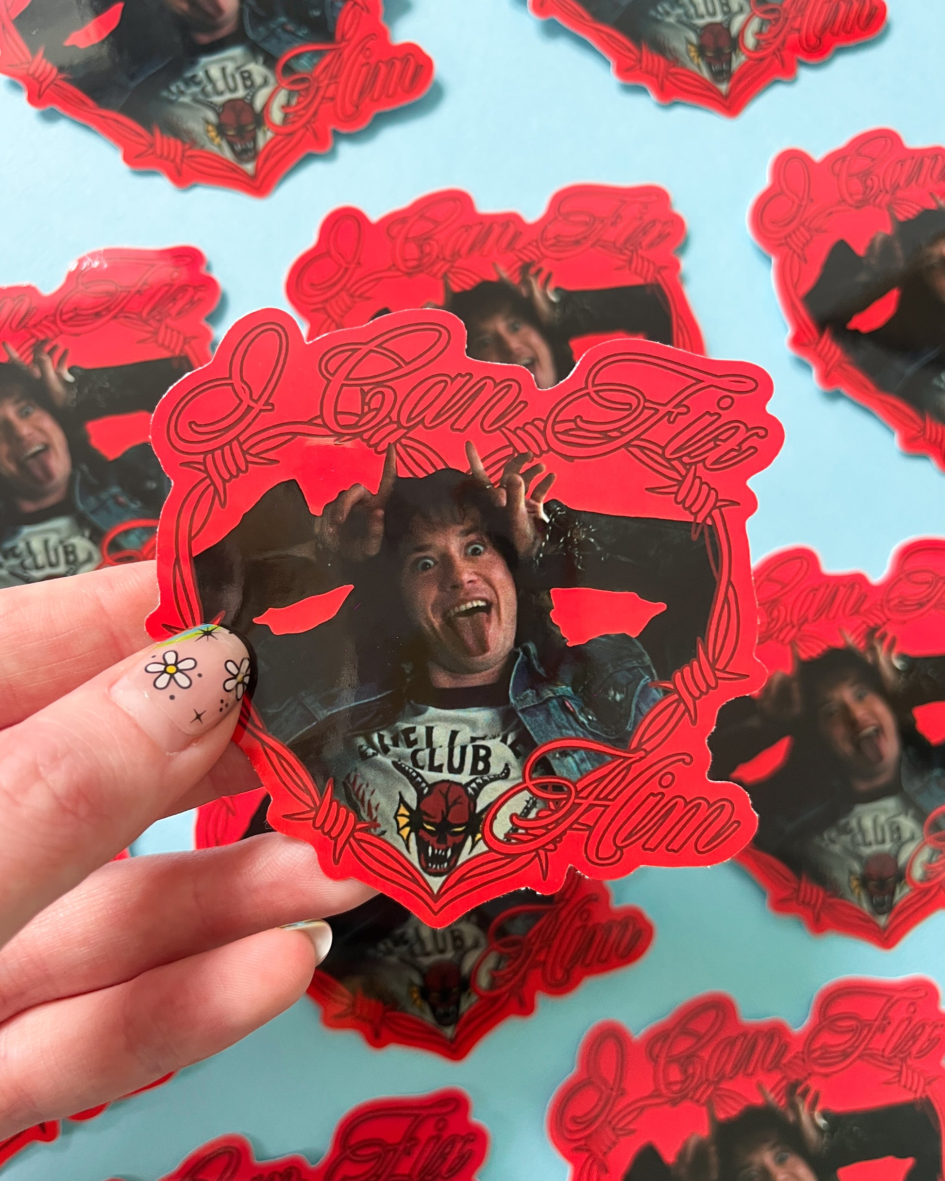 A person holding a vinyl sticker with a holographic finish featuring the character Eddie Munson, with red and black colors predominately.