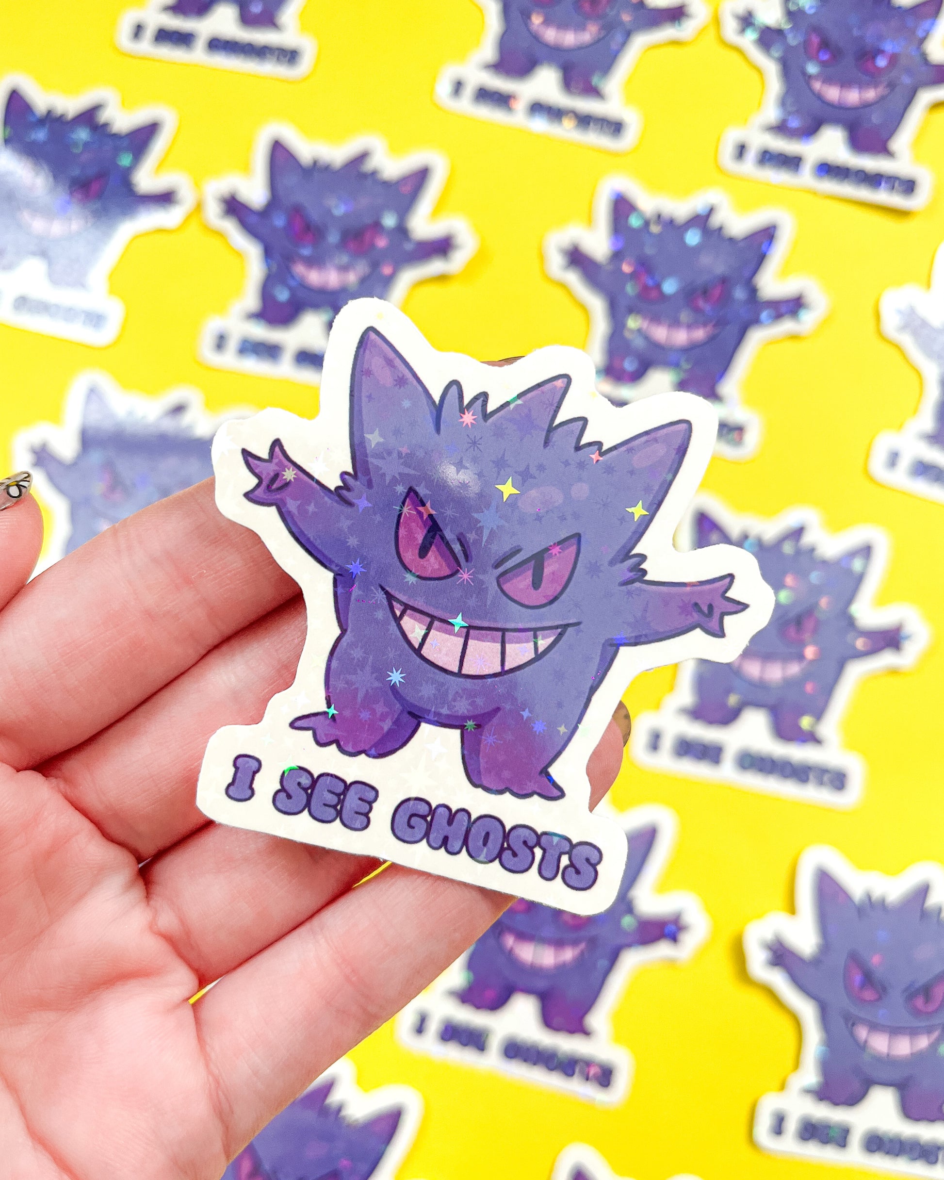 A holographic vinyl sticker of Gengar Pokemon with a caption 'I SEE GHOSTS' written at the bottom. The sticker is held against a background with more similar stickers.