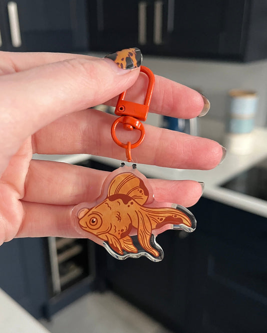 A hand holding a goldfish-shaped keyring with an orange swivel clasp.