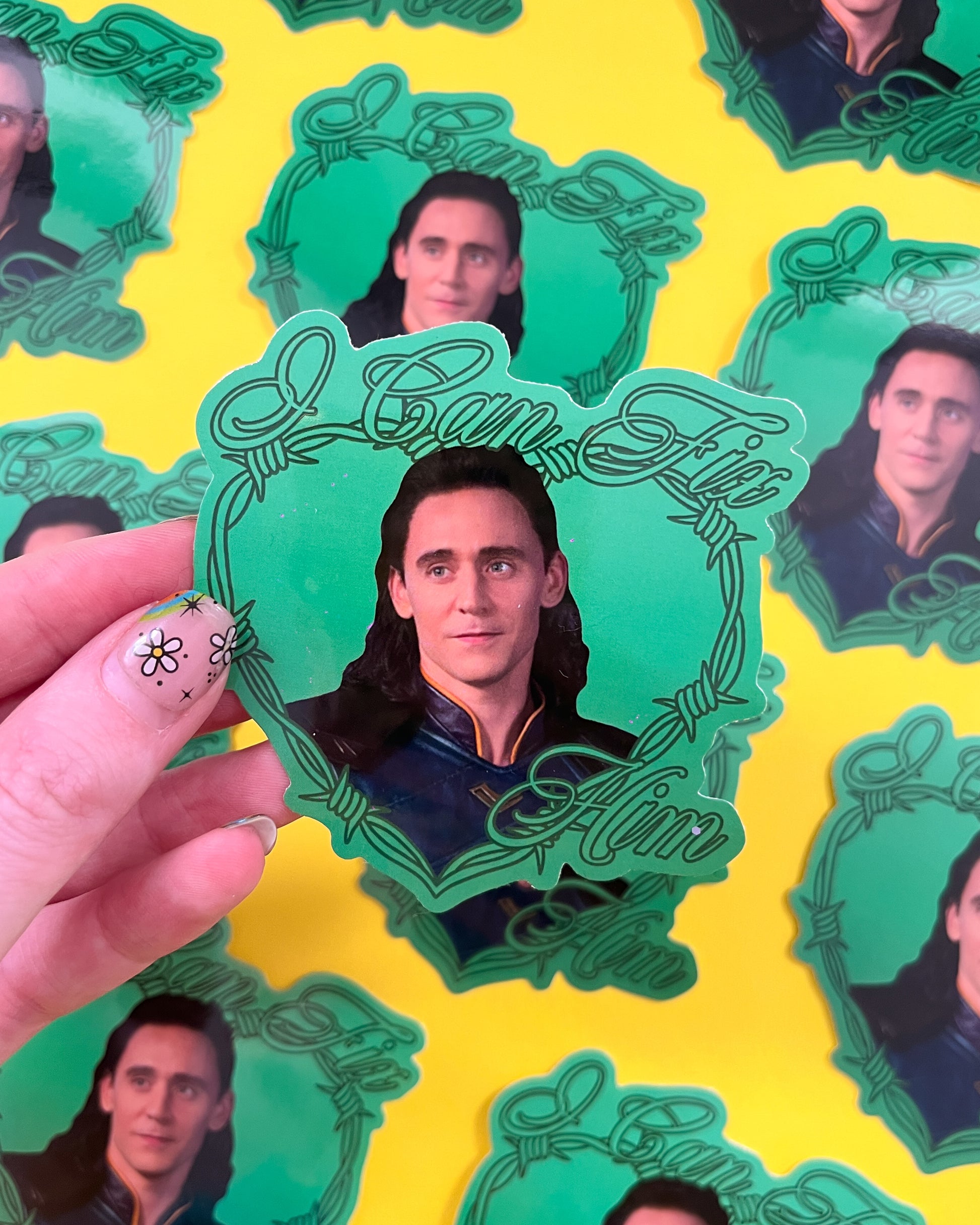 A person holding a vinyl sticker with an image of Loki from Marvel, with multiple stickers displayed in the background.