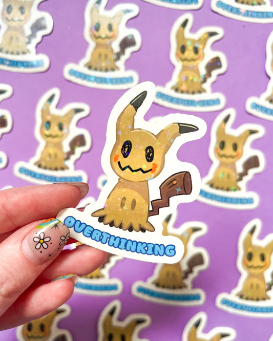 A holographic vinyl sticker of Mimikyu, a Pokemon character, held against a background of the same sticker pattern, displaying a shiny finish.