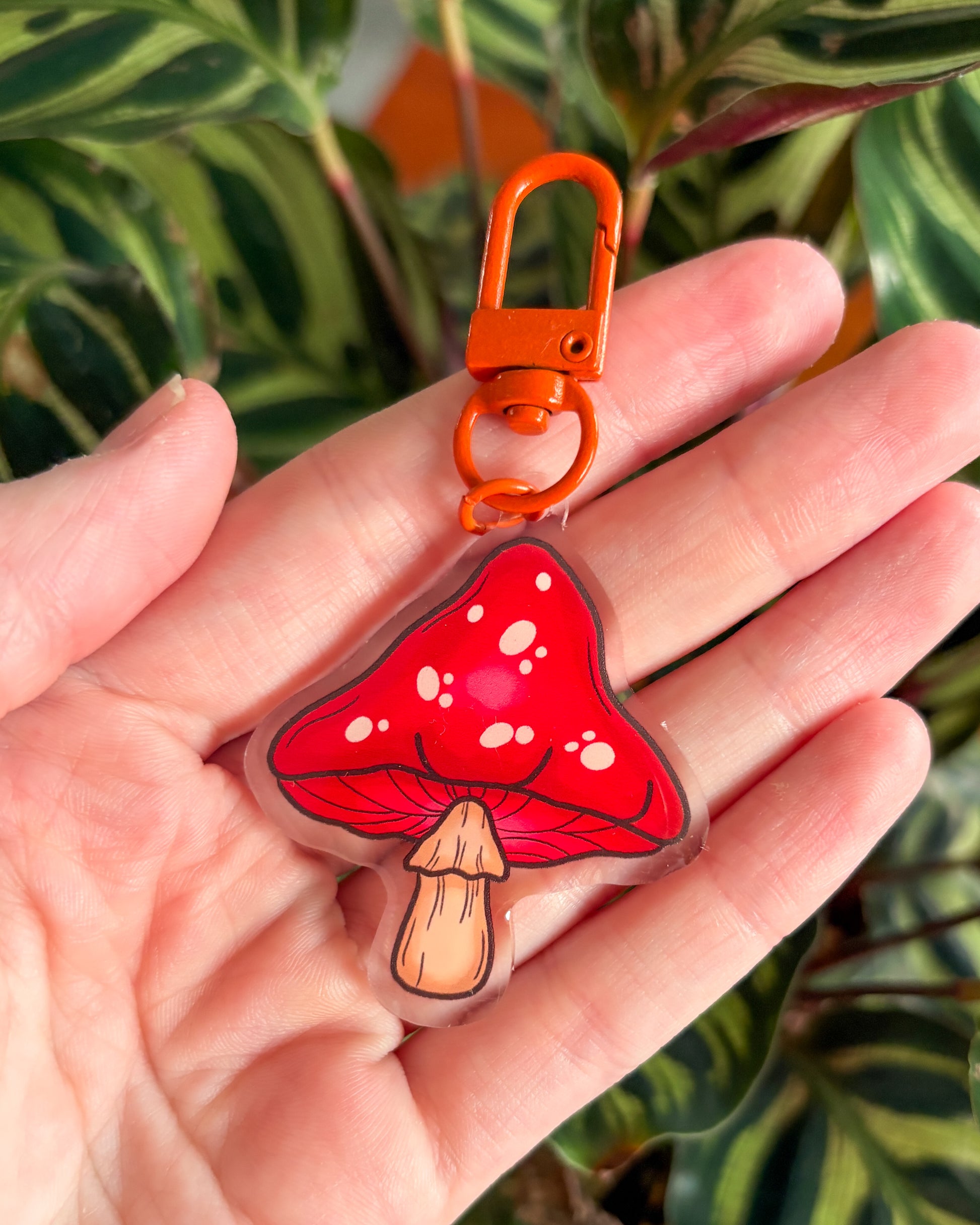 Red mushroom-shaped keychain with an orange clip held in a hand against a plant background