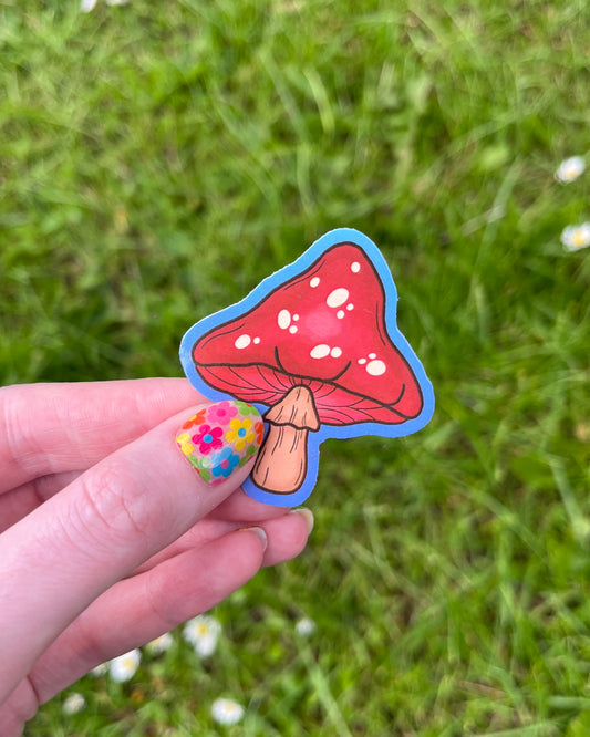 A hand holding a red and white mushroom-shaped vinyl sticker with a grassy background.
