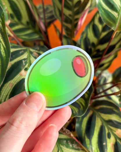 Hand holding a green olive sticker in front of a plant