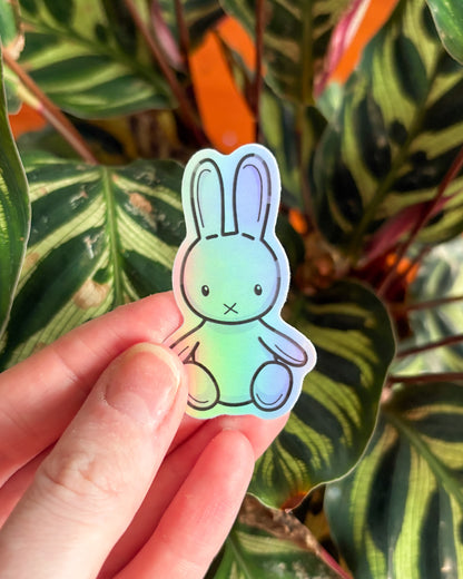 Hand holding a pastel-colored bunny sticker in front of a leafy green plant