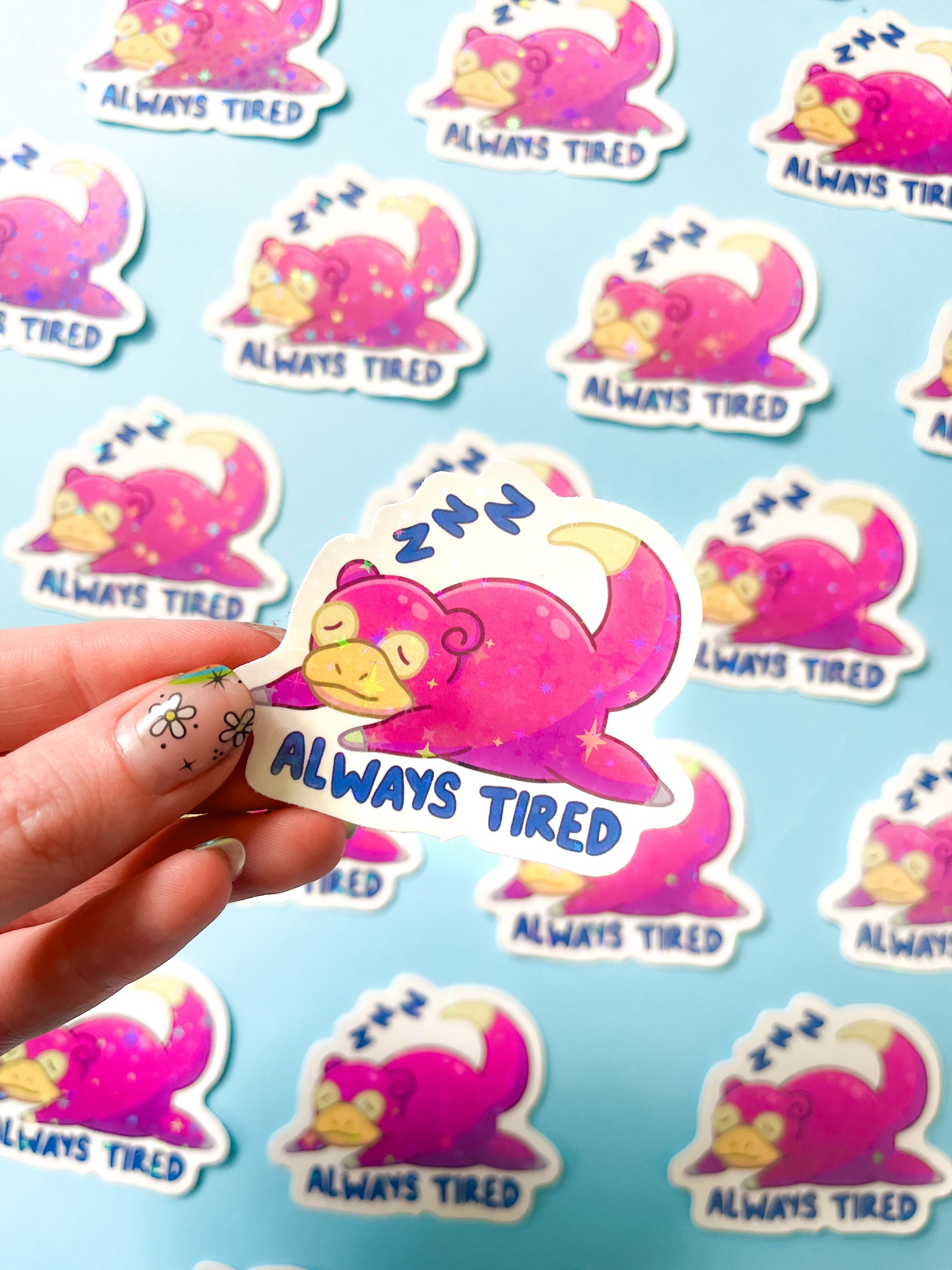 A person holding a vinyl sticker of Slowpoke Pokemon with the text 'ALWAYS TIRED' on it, background is filled with similar stickers creating a pattern.