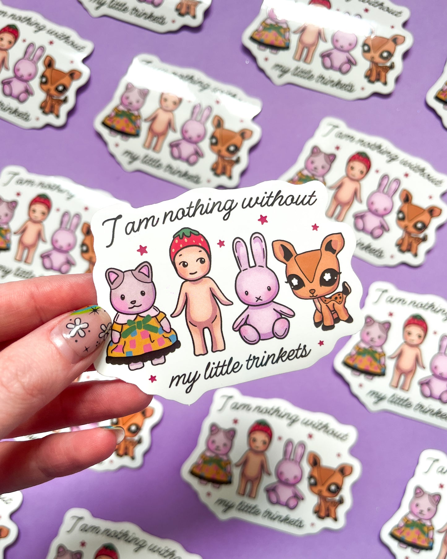 A vinyl sticker with an illustration of a cartoonish character and a rabbit-like creature, accompanied by the phrase 'I am nothing without my little trinkets'. The sticker is held against a purple background with similar stickers scattered around.