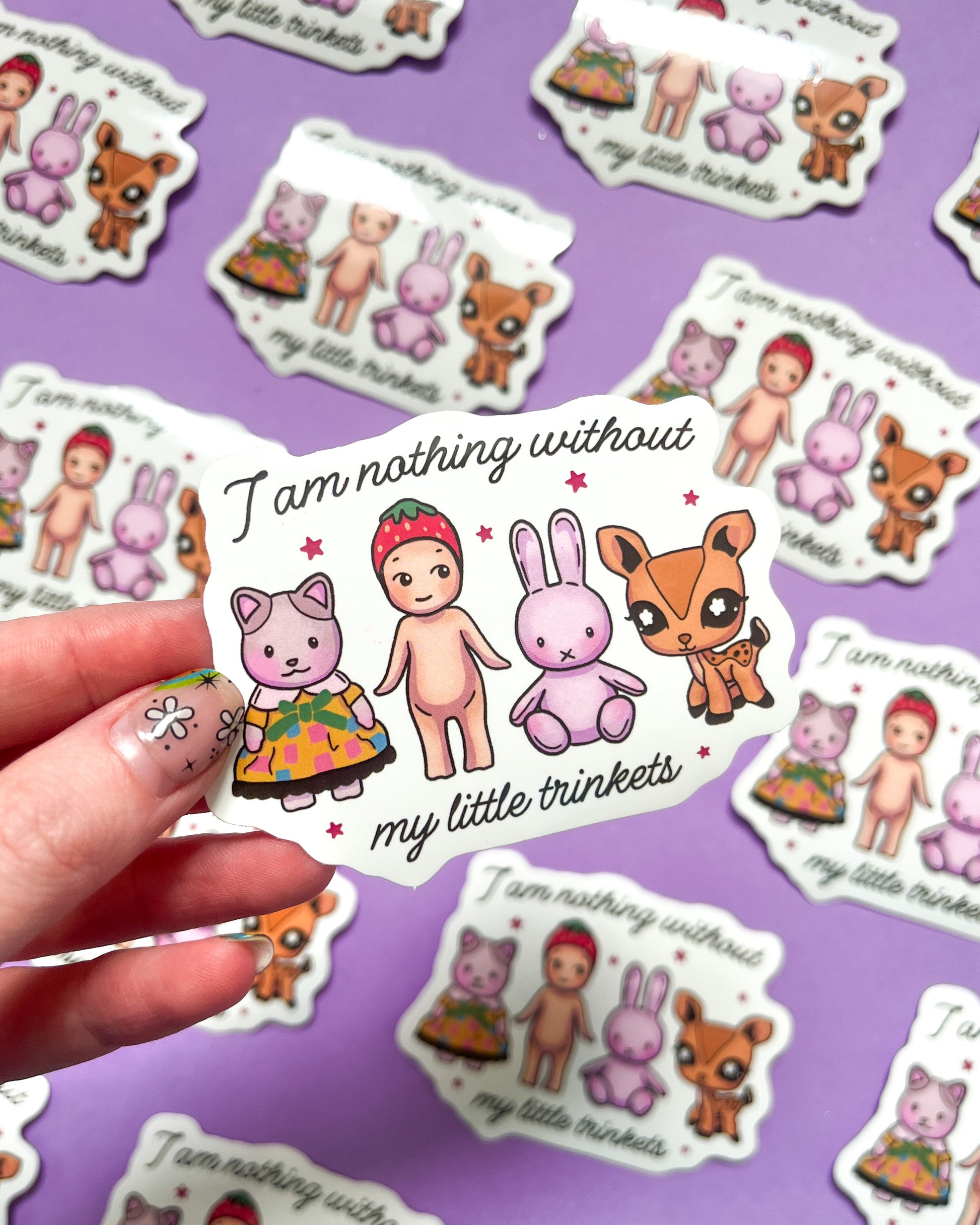 A vinyl sticker with an illustration of a cartoonish character and a rabbit-like creature, accompanied by the phrase 'I am nothing without my little trinkets'. The sticker is held against a purple background with similar stickers scattered around.
