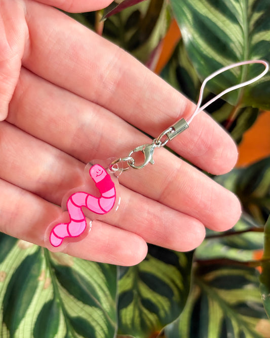 Pink worm charm on a clear plastic holder held between fingers with a green leafy background.